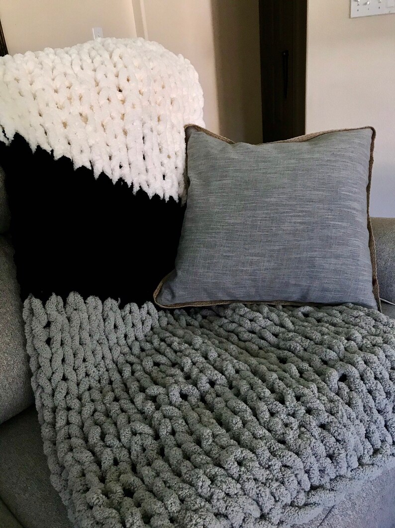 May include: A gray chunky knit throw blanket with a white and black patterned throw pillow and a gray throw pillow with a brown border on a gray couch.