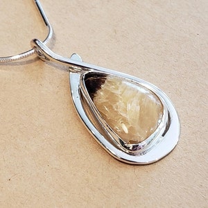 May include: A silver-toned pendant necklace featuring a teardrop-shaped, translucent, light-brown and cream-colored stone set within a polished, curved metal frame. The pendant hangs from a thin, flexible silver-toned chain with a smooth finish.