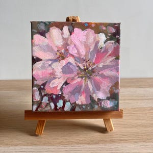 May include: A small square canvas painting featuring an impressionistic floral design with layered brushstrokes in shades of pink, white, and deep brown. The artwork is displayed on a miniature wooden easel, showcasing a textured paint application.