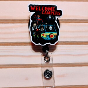May include: A retractable badge reel with a horror-themed design. The image features a black and white graphic of a van with characters from horror movies. The text "WELCOME CAMPERS" is in red letters.