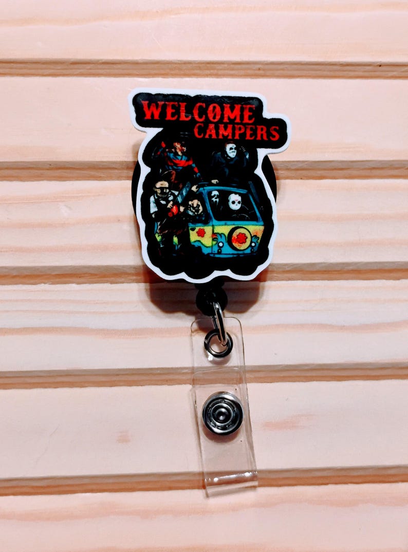 May include: A retractable badge reel with a horror-themed design. The image features a black and white graphic of a van with characters from horror movies. The text "WELCOME CAMPERS" is in red letters.