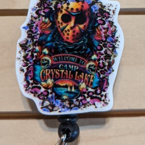 May include: A badge reel featuring a colorful design with a horror theme. The image includes a hockey mask, floral accents, and the text "WELCOME TO CAMP CRYSTAL LAKE". The reel has a black body and a clear plastic clip.
