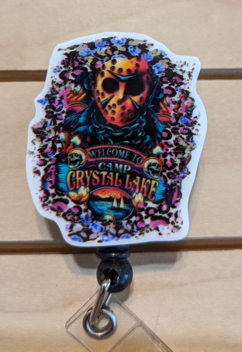 May include: A badge reel featuring a colorful design with a horror theme. The image includes a hockey mask, floral accents, and the text "WELCOME TO CAMP CRYSTAL LAKE". The reel has a black body and a clear plastic clip.
