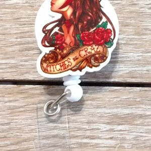 May include: A white plastic retractable badge holder with a red, white, and gold cartoon image of a woman with red hair and a red headband. The image has the text "Witches Be Crazy" on a banner.