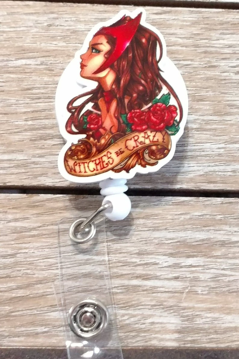 May include: A white plastic retractable badge holder with a red, white, and gold cartoon image of a woman with red hair and a red headband. The image has the text "Witches Be Crazy" on a banner.