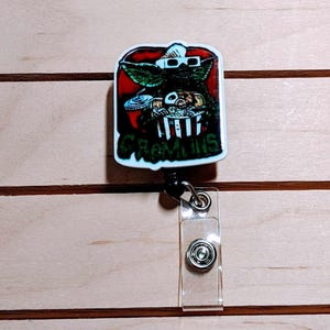 May include: A rectangular badge reel featuring a Gremlin character wearing 3D glasses, holding popcorn and a drink, with the word "Gremlins" below. The design is set against a red background with a white border. The reel has a clear plastic strap and a metal snap.