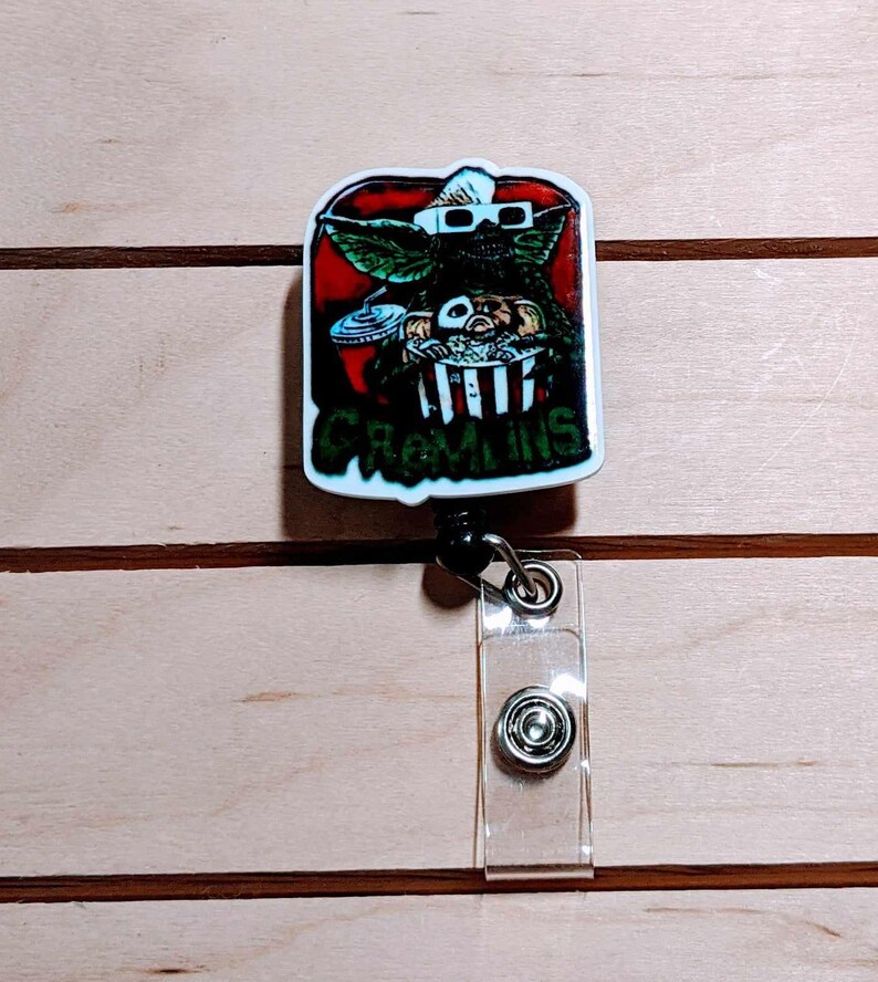 May include: A rectangular badge reel featuring a Gremlin character wearing 3D glasses, holding popcorn and a drink, with the word "Gremlins" below. The design is set against a red background with a white border. The reel has a clear plastic strap and a metal snap.