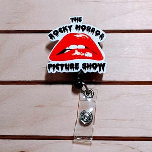 May include: A retractable badge reel featuring the iconic red lips logo from "The Rocky Horror Picture Show." The design is set against a white background with black text. The reel has a clear plastic strap and a black cord.