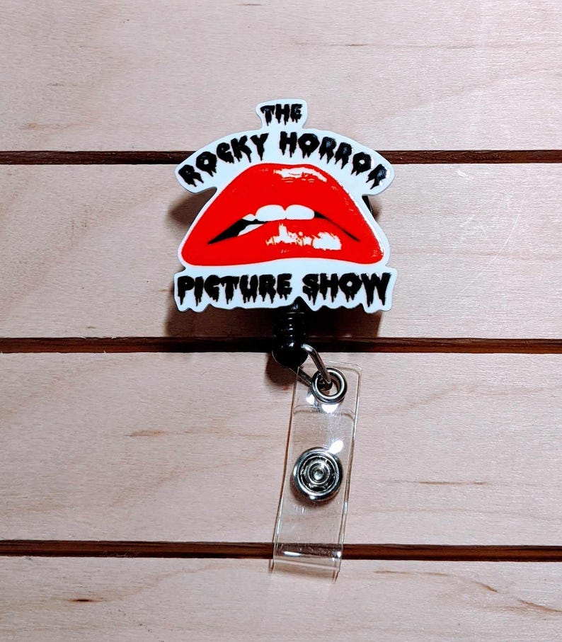 May include: A retractable badge reel featuring the iconic red lips logo from "The Rocky Horror Picture Show." The design is set against a white background with black text. The reel has a clear plastic strap and a black cord.