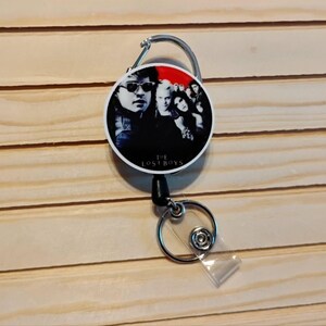 May include: A retractable badge reel featuring a group of people with sunglasses and dark clothing against a red and black background. The text "THE LOST BOYS" is printed at the bottom of the reel. The reel has a silver carabiner clip and a clear plastic strap.
