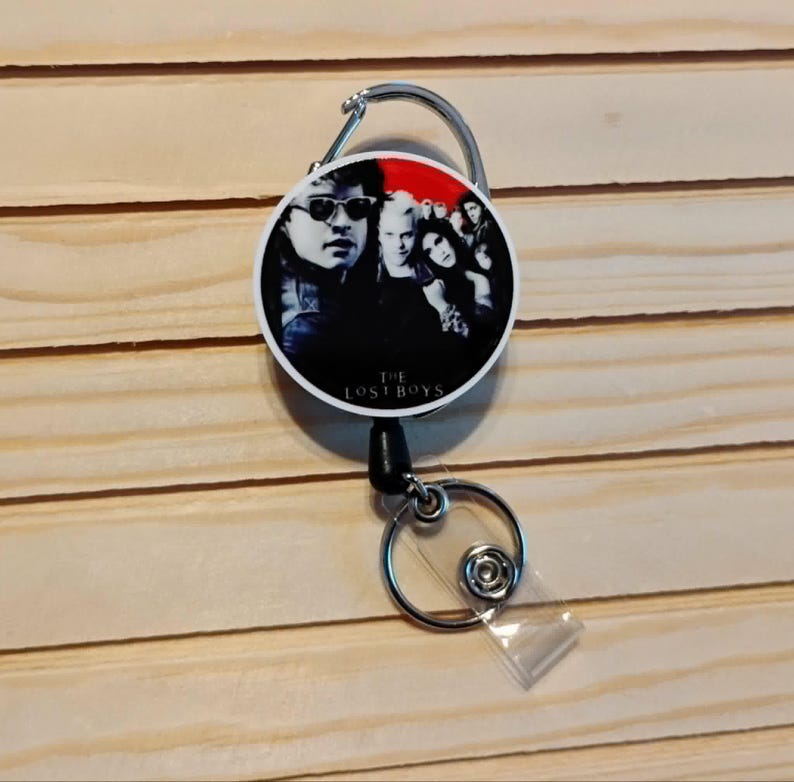 May include: A retractable badge reel featuring a group of people with sunglasses and dark clothing against a red and black background. The text "THE LOST BOYS" is printed at the bottom of the reel. The reel has a silver carabiner clip and a clear plastic strap.