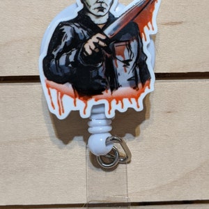 May include: A white retractable badge reel featuring a graphic of Michael Myers from the Halloween horror film series. The image shows Myers holding a bloody knife. The reel has a white plastic body and a metal clip.