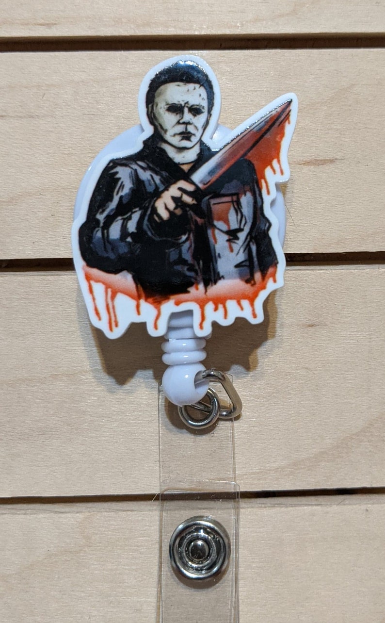 May include: A white retractable badge reel featuring a graphic of Michael Myers from the Halloween horror film series. The image shows Myers holding a bloody knife. The reel has a white plastic body and a metal clip.