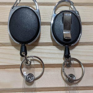 May include: Two black retractable badge holders with silver carabiner clips and key rings. The badge reels have a textured surface and a clear plastic strap for attaching an ID badge. The background is a light wood.