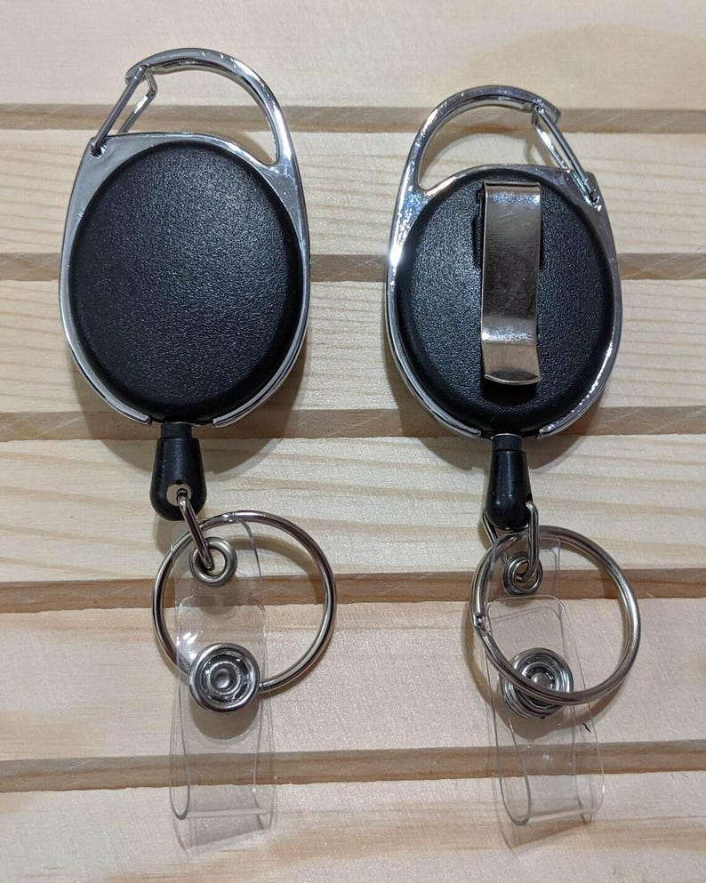 May include: Two black retractable badge holders with silver carabiner clips and key rings. The badge reels have a textured surface and a clear plastic strap for attaching an ID badge. The background is a light wood.