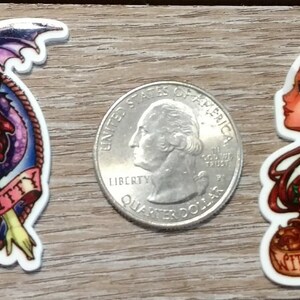 May include: Two colorful cartoon character stickers. The first sticker features a pink-haired woman with a purple dragon, with the text "Hello Kitty". The second sticker features a red-haired woman with a red crown, with the text "Witches be crazy".