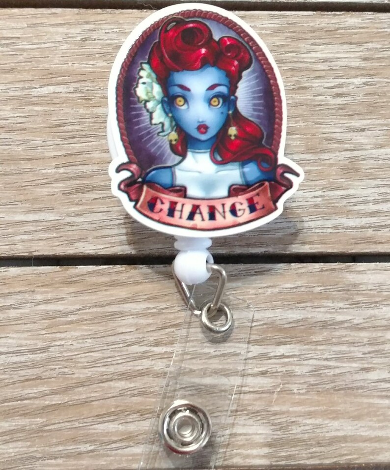 May include: A white retractable badge holder with a round, colorful graphic featuring a woman with red hair and blue skin. The graphic has the word "CHANGE" written on a banner.