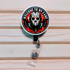May include: A round, white badge reel with the text "WELCOME TO MY CAMP" encircling a graphic of a hockey mask and a figure in a black jacket against a red and orange background. The badge reel has a black retractable cord and a clear plastic strap.