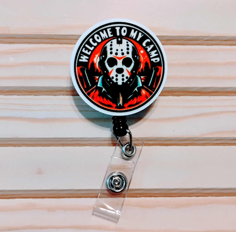 May include: A round, white badge reel with the text "WELCOME TO MY CAMP" encircling a graphic of a hockey mask and a figure in a black jacket against a red and orange background. The badge reel has a black retractable cord and a clear plastic strap.