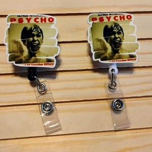 May include: Two white badge reels featuring the movie poster for Alfred Hitchcock's "Psycho." The reel design includes the movie title in red and the image of a screaming man. Each reel has a clear plastic strap and a metal clip.