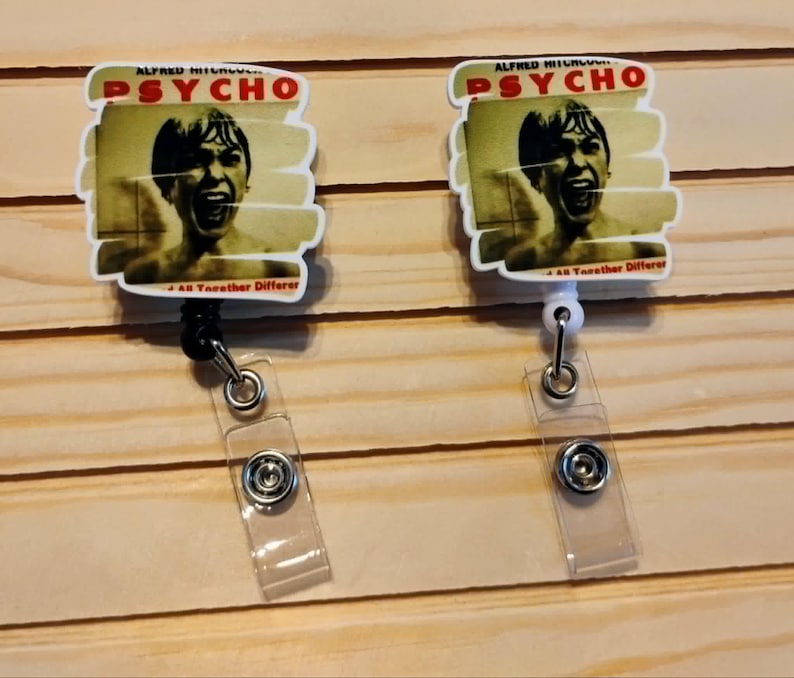 May include: Two white badge reels featuring the movie poster for Alfred Hitchcock's "Psycho." The reel design includes the movie title in red and the image of a screaming man. Each reel has a clear plastic strap and a metal clip.
