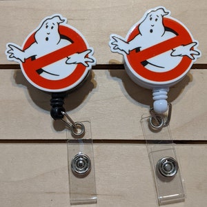 May include: Two badge reels featuring the Ghostbusters logo: a white ghost inside a red circle with a red slash. The reels have a black and white design, with a clear plastic strap and a metal snap closure.