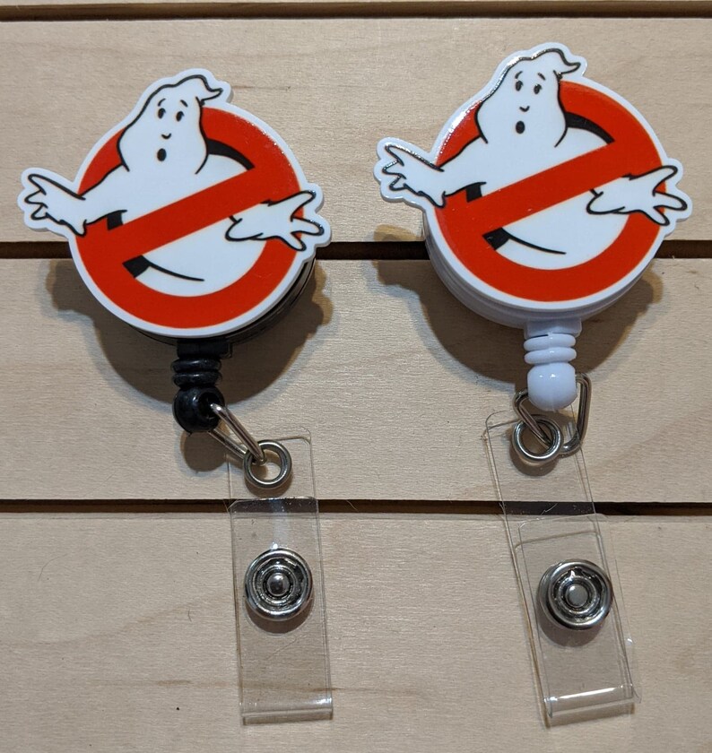 May include: Two badge reels featuring the Ghostbusters logo: a white ghost inside a red circle with a red slash. The reels have a black and white design, with a clear plastic strap and a metal snap closure.