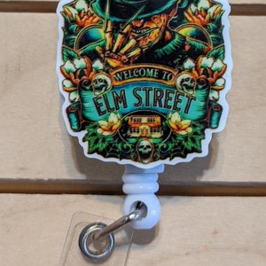 May include: A badge reel with a colorful graphic featuring Freddy Krueger from the movie "A Nightmare on Elm Street." The design includes the text "Welcome to Elm Street" and floral accents. The reel is white with a clear plastic strap.