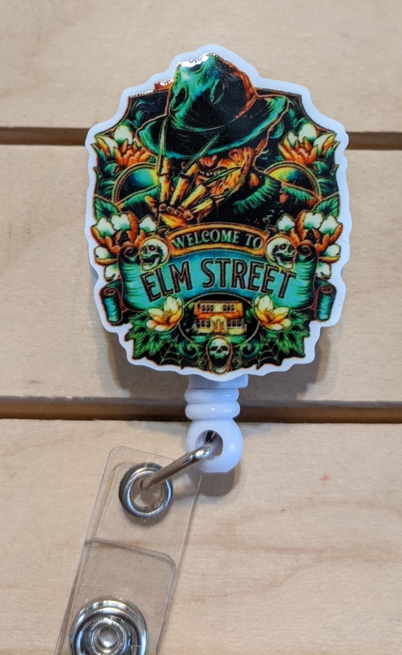 May include: A badge reel with a colorful graphic featuring Freddy Krueger from the movie "A Nightmare on Elm Street." The design includes the text "Welcome to Elm Street" and floral accents. The reel is white with a clear plastic strap.