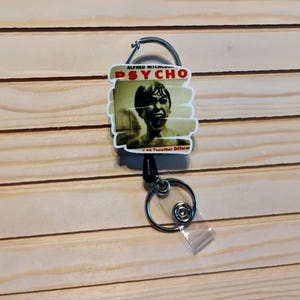 May include: A badge reel featuring a graphic from the movie "Psycho." The reel has a silver clip and a black retractable cord. The graphic includes the movie title and a still image of a person screaming.