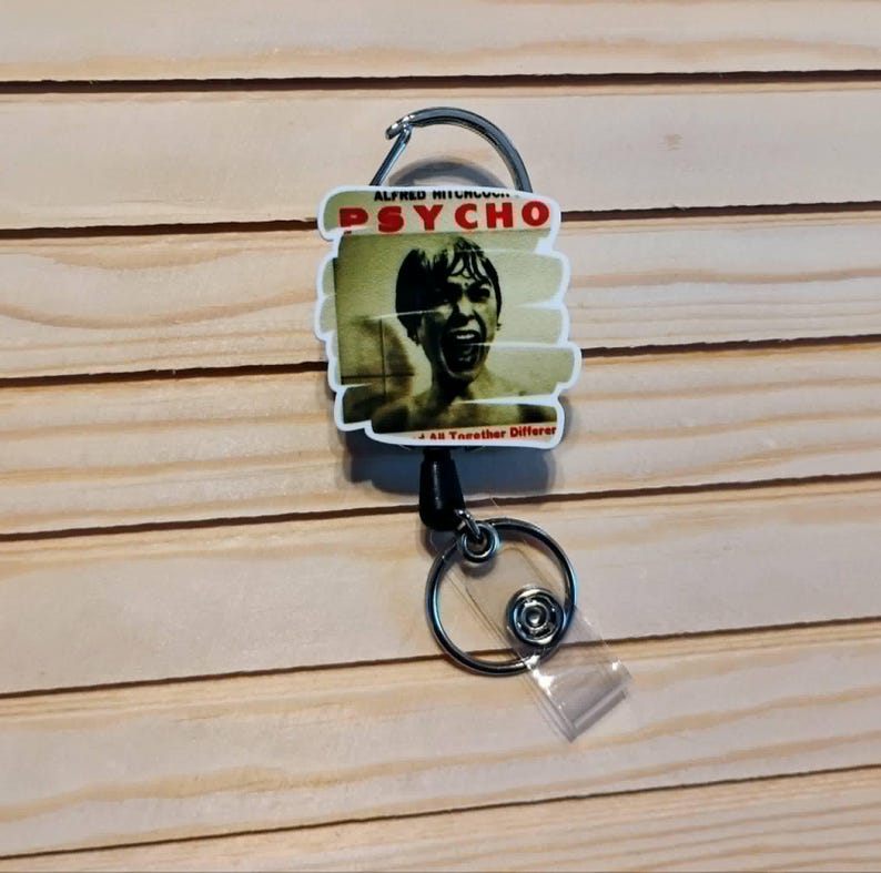 May include: A badge reel featuring a graphic from the movie "Psycho." The reel has a silver clip and a black retractable cord. The graphic includes the movie title and a still image of a person screaming.