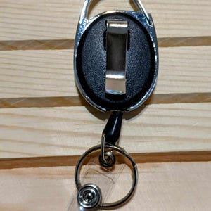 May include: A retractable badge reel with a silver carabiner clip, black face, and a clear plastic strap. The reel has a silver metal ring and a black cord. The background is a light brown wooden surface.