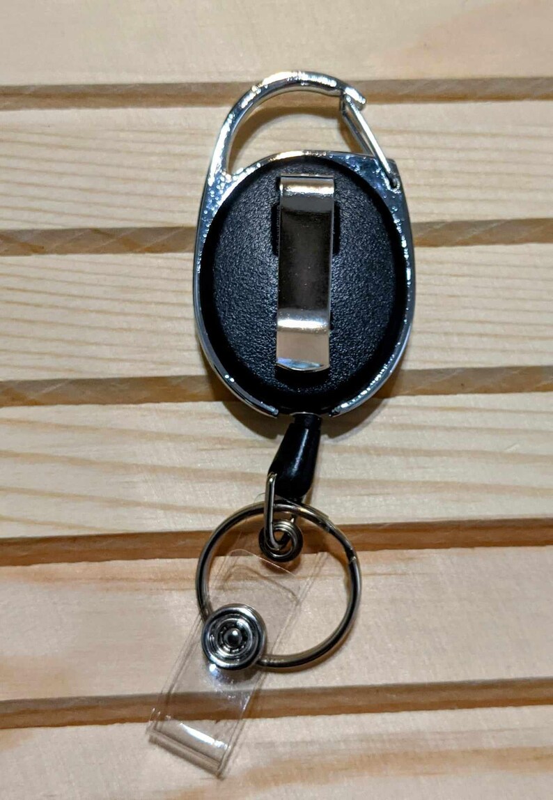 May include: A retractable badge reel with a silver carabiner clip, black face, and a clear plastic strap. The reel has a silver metal ring and a black cord. The background is a light brown wooden surface.