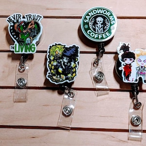 May include: Four retractable badge reels with cartoon character designs. One reel says "Never Trust The Living" with a green and black graphic. Another features a "Sandworms Coffee" logo. The other two reels have cartoon characters.
