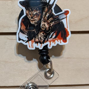 May include: A retractable badge reel featuring a graphic of a horror movie character with a fedora and bladed glove. The image has a white border and red blood-like drips. The reel has a black cord and a clear plastic badge holder.