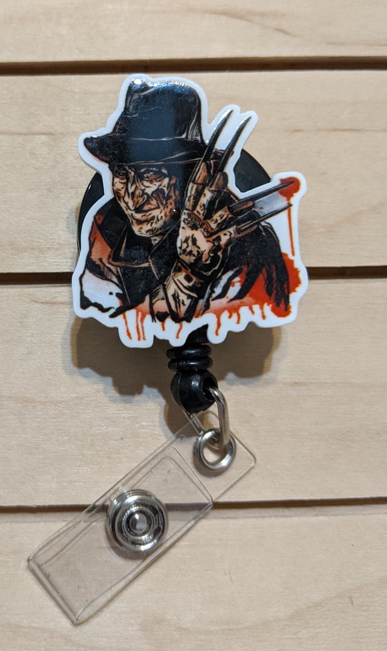 May include: A retractable badge reel featuring a graphic of a horror movie character with a fedora and bladed glove. The image has a white border and red blood-like drips. The reel has a black cord and a clear plastic badge holder.