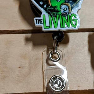 May include: A retractable badge reel with a graphic design featuring the text "NEVER TRUST THE LIVING". The design includes a green and red color scheme with a zombie-like character. The reel has a clear plastic strap.