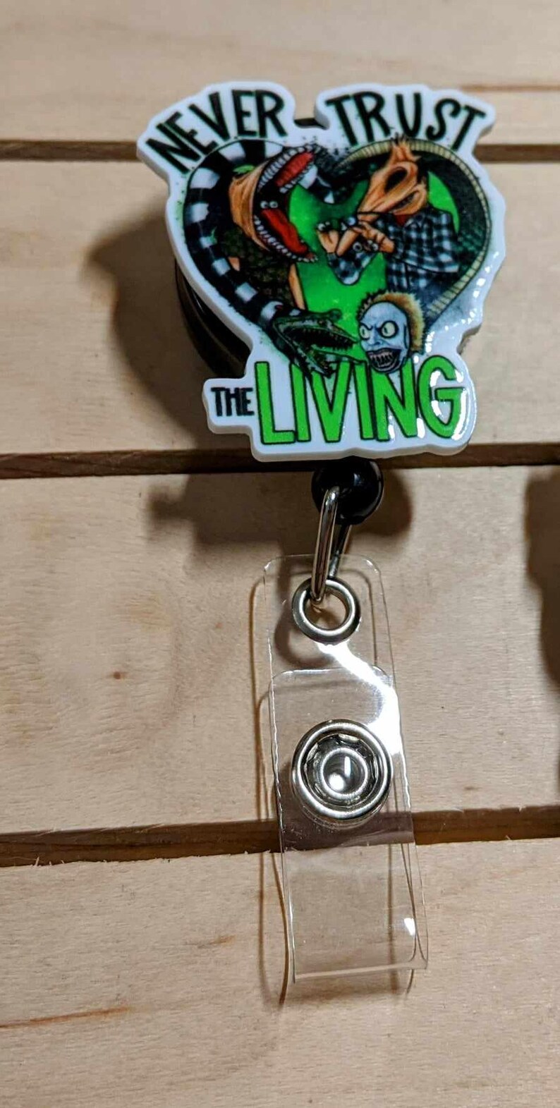 May include: A retractable badge reel with a graphic design featuring the text "NEVER TRUST THE LIVING". The design includes a green and red color scheme with a zombie-like character. The reel has a clear plastic strap.