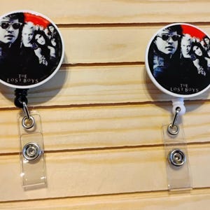 May include: Two white and black badge reels with a circular design featuring a group of people, with the text "THE LOST BOYS". The reels have a clear plastic strap and a metal clip.