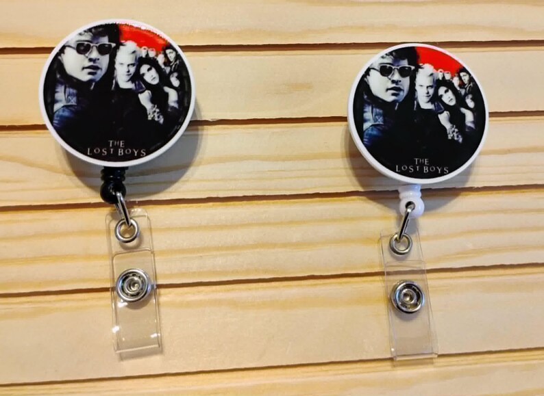 May include: Two white and black badge reels with a circular design featuring a group of people, with the text "THE LOST BOYS". The reels have a clear plastic strap and a metal clip.