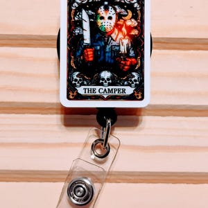 May include: A retractable badge reel featuring a horror-themed tarot card design. The card depicts a masked figure holding a knife and a candle, with the text "THE CAMPER" at the bottom. The reel has a clear plastic strap and a metal clip.