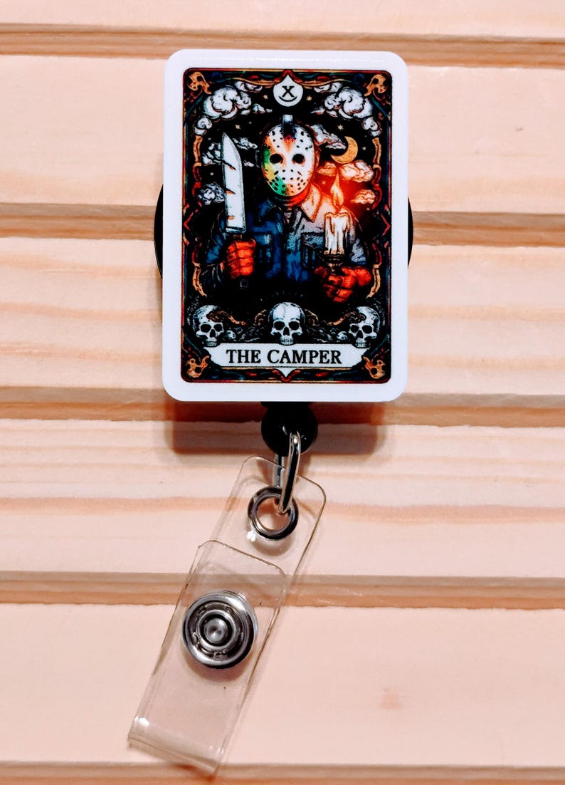 May include: A retractable badge reel featuring a horror-themed tarot card design. The card depicts a masked figure holding a knife and a candle, with the text "THE CAMPER" at the bottom. The reel has a clear plastic strap and a metal clip.