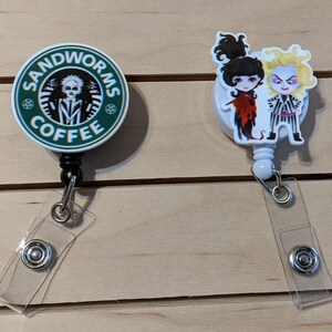 May include: Two retractable badge holders. One features a green and white design with the text "SANDWORMS COFFEE" and a cartoon character. The other has cartoon characters in red, black, and white. Both have clear plastic straps and metal snaps.