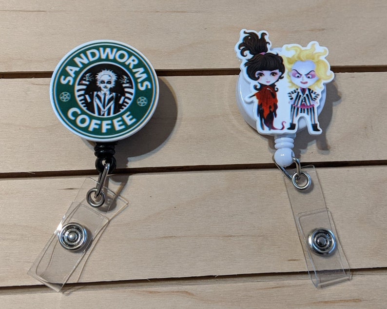 May include: Two retractable badge holders. One features a green and white design with the text "SANDWORMS COFFEE" and a cartoon character. The other has cartoon characters in red, black, and white. Both have clear plastic straps and metal snaps.