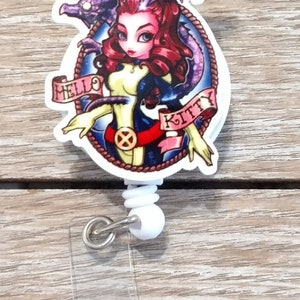 May include: A white retractable badge holder with a round, colorful graphic featuring a cartoon woman with red hair, a blue and yellow outfit, and a purple dragon. The graphic has the text "Hello Kitty" on a banner.