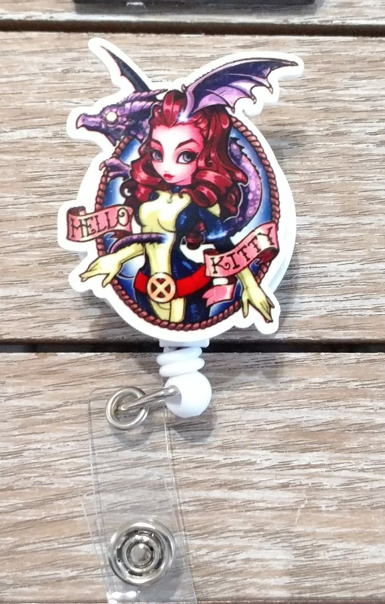 May include: A white retractable badge holder with a round, colorful graphic featuring a cartoon woman with red hair, a blue and yellow outfit, and a purple dragon. The graphic has the text "Hello Kitty" on a banner.