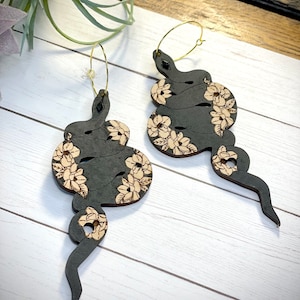 Peut inclure: Une paire de boucles d'oreilles en forme de serpent noir avec des détails floraux. Les boucles d'oreilles sont en bois et ont des anneaux dorés.