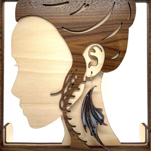 Pu&ograve; includere: Un espositore per orecchini in legno con un ritaglio di una testa di persona. Un singolo orecchino a forma di ala di drago nero e argento &egrave; esposto sullo stand.
