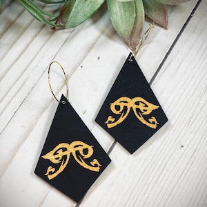 May include: A pair of black diamond-shaped earrings with gold-colored bow designs. The earrings hang from gold-colored hoop wires. The earrings are displayed on a white wooden surface with a succulent plant in the background.