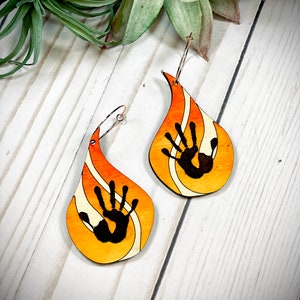 May include: A pair of teardrop-shaped wooden earrings with a handprint design. The earrings are painted in shades of orange, yellow, and white, with a black handprint in the centre. The earrings are attached to silver hoops.